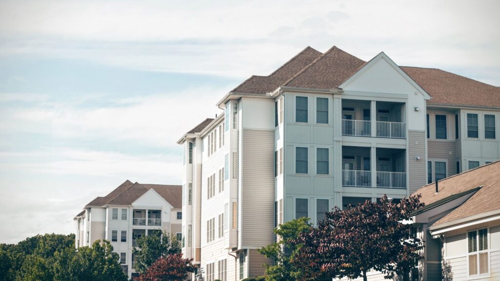 Modern apartment buildings with light-colored siding and multiple balconies, set against a partly cloudy sky. There are green trees and reddish-leaved trees in front of the buildings.