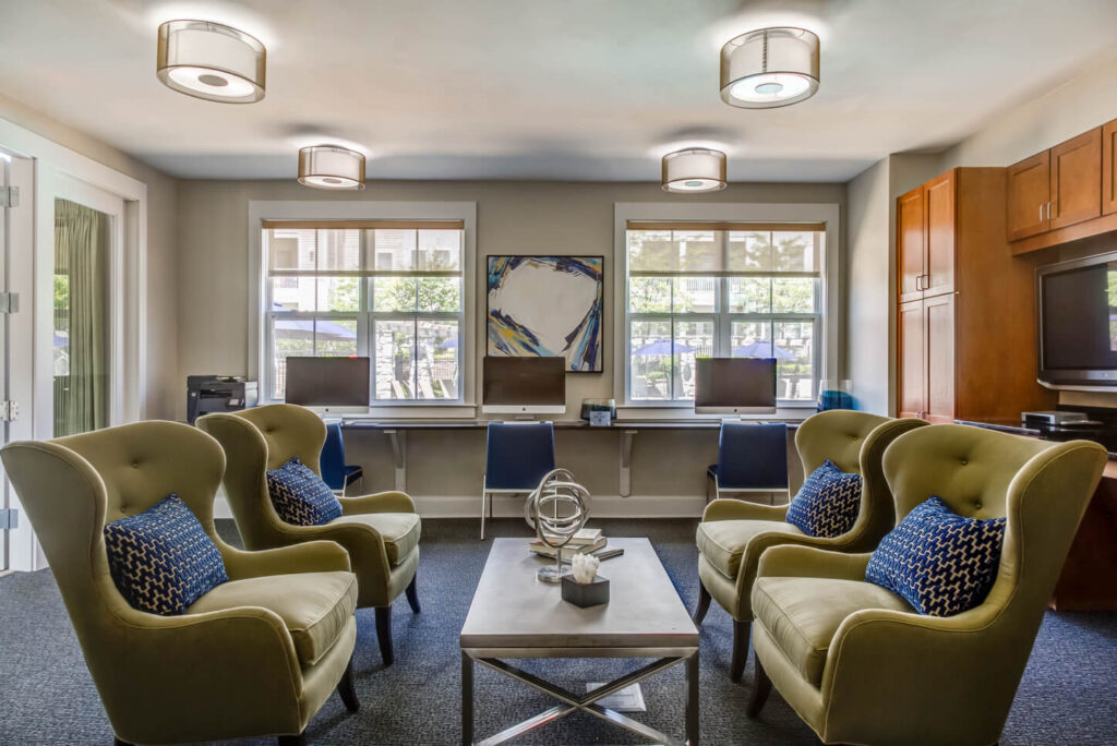 Modern lounge with four green armchairs, blue pillows, a coffee table, and a work area with computers along the back wall under large windows. Contemporary lighting and wall art complete the space.