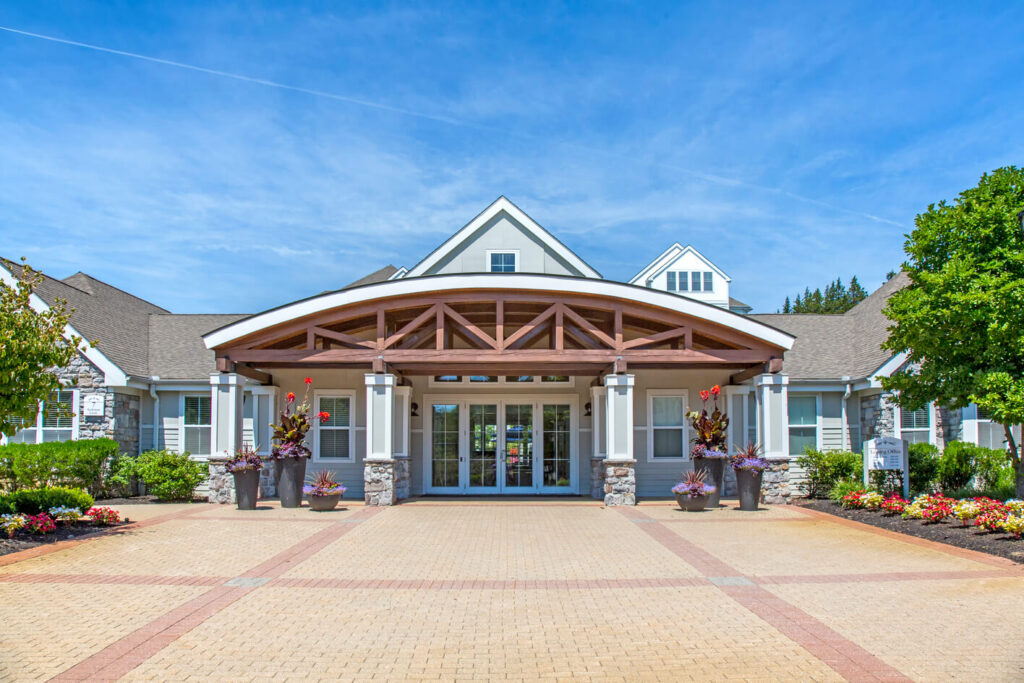 A modern building with a large wooden entrance canopy, stone columns, potted plants, and glass doors, surrounded by landscaped flower beds on a sunny day.
