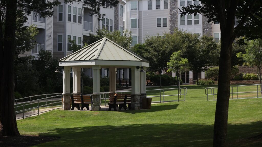 A small gazebo with benches sits on a grassy lawn, surrounded by trees and modern apartment buildings in the background on a sunny day.