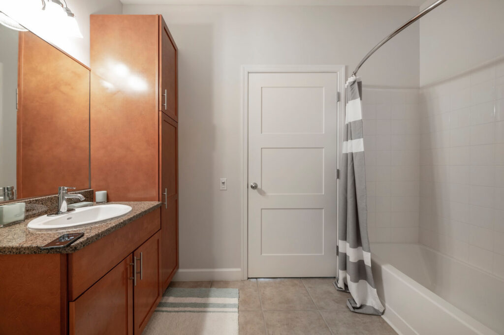 Modern bathroom with a wood vanity, granite countertop, sink, mirror, and tall cabinet. Theres a bathtub with a striped gray and white shower curtain and a white door in the background.