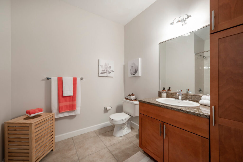 Modern bathroom with light beige walls, wood cabinets, granite countertop, a large mirror, white toilet, towel rack with coral and white towels, two floral wall artworks, and tile flooring.