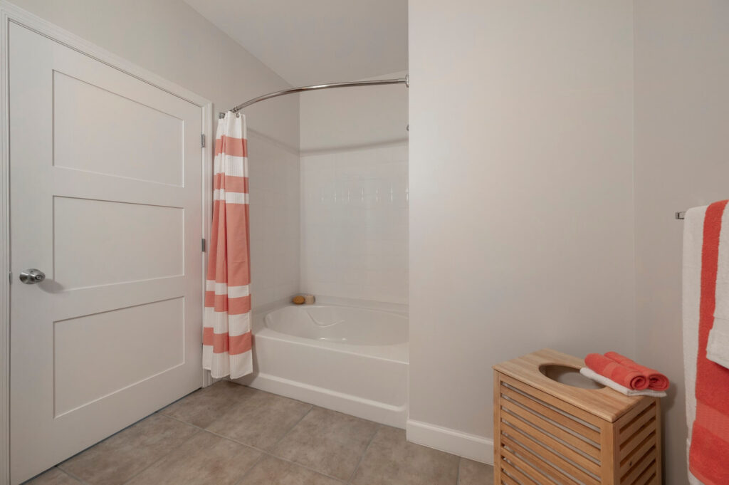 A bathroom with a white bathtub, coral and white striped shower curtain, a wooden laundry hamper, gray tile floor, and white walls. Two coral and white towels hang on a rack beside the tub.