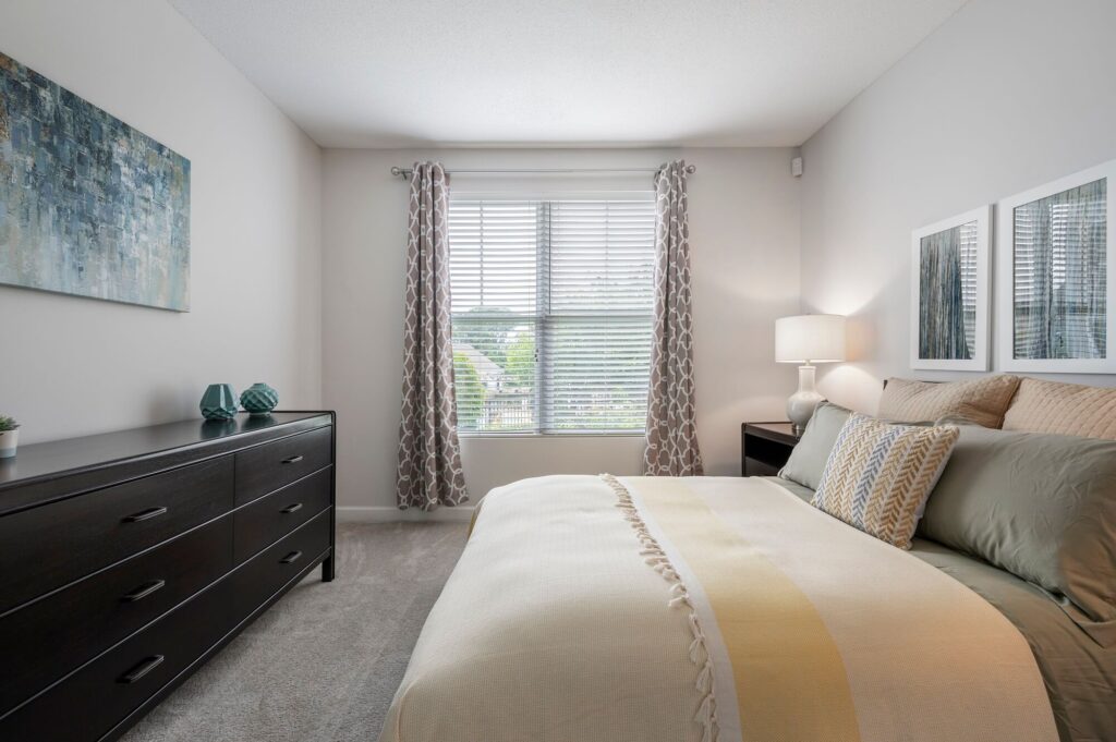 A modern bedroom with a neatly made bed, two nightstands, a dresser, abstract wall art, and a window with patterned curtains letting in natural light.