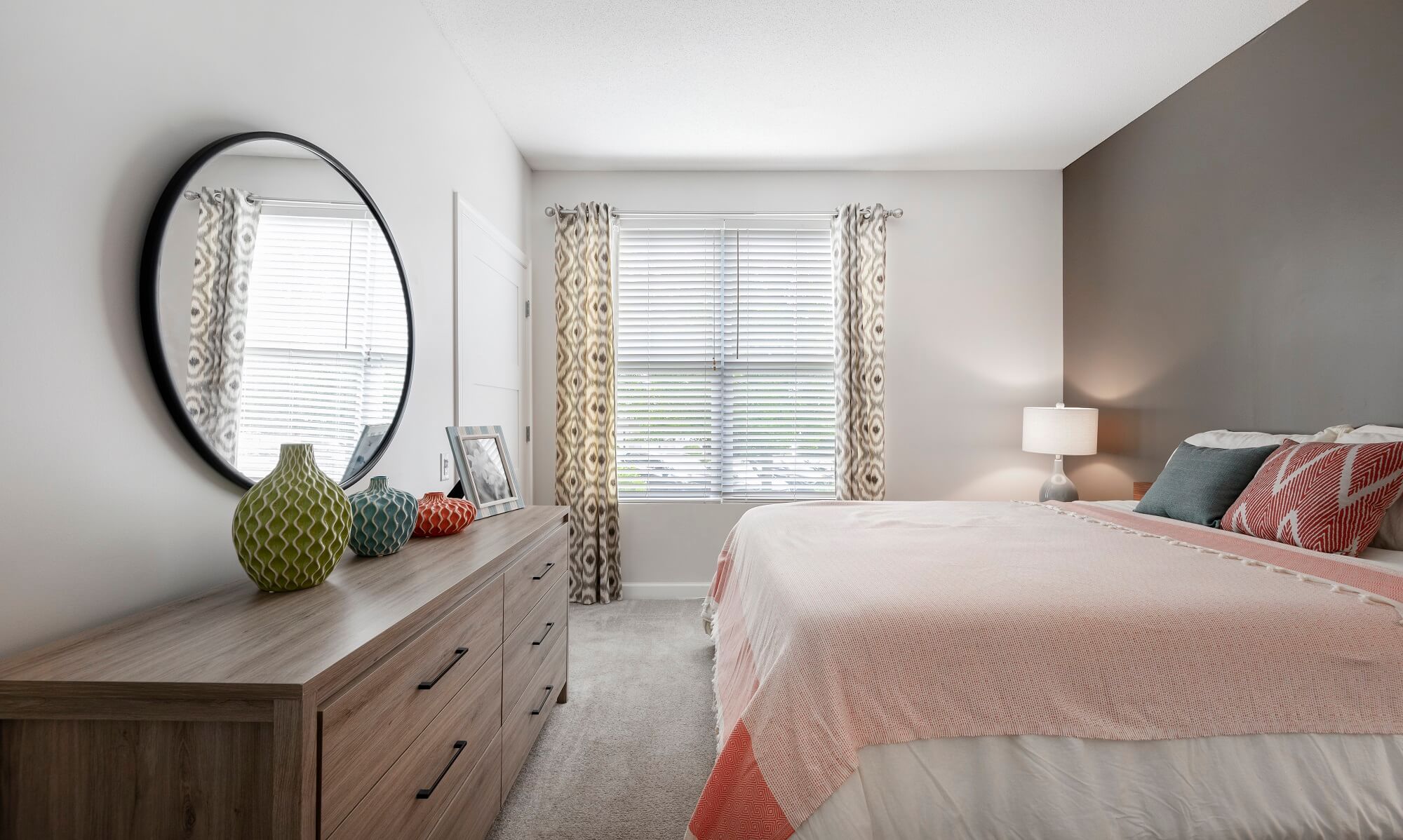 Modern bedroom with a wood dresser, round mirror, decorative vases, bed with pink bedding, patterned pillows, a window with curtains, and a lamp on a nightstand.
