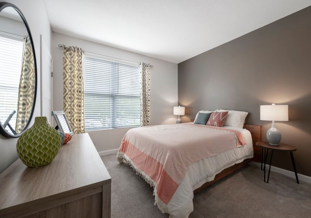 Modern bedroom with a large window, bed with pink and white bedding, two bedside tables with lamps, a dresser with a green vase, and a round mirror on the wall. Light gray carpet and neutral walls.
