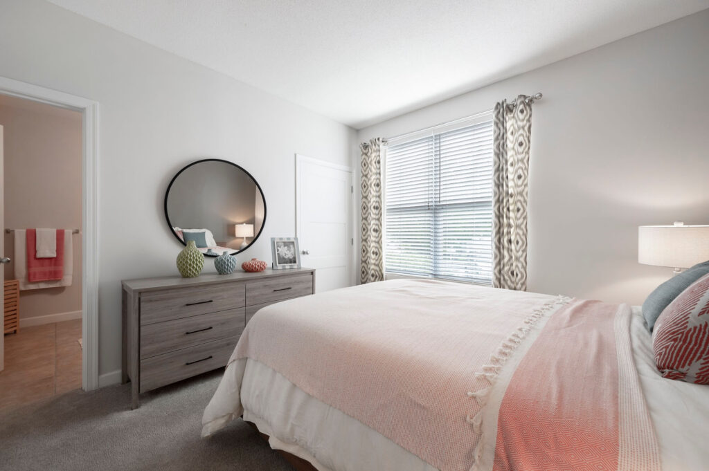 A modern bedroom with a bed, gray dresser, round mirror, decorative vases, a lamp, and a window with patterned curtains letting in natural light. A bathroom is visible through an open door.
