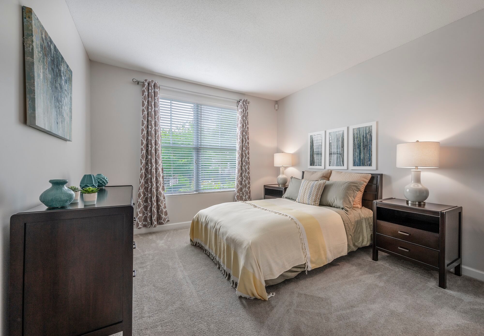 A neatly made bed with beige bedding sits between two dark wood nightstands with lamps. Large window with patterned curtains lets in light. Art decorates light gray walls. Carpeted floor.