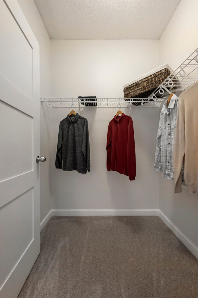 A walk-in closet with beige carpet, white walls, and white wire shelves. Several shirts hang on hangers, and there are baskets and a black box on the shelves. The closet door is partially visible on the left.