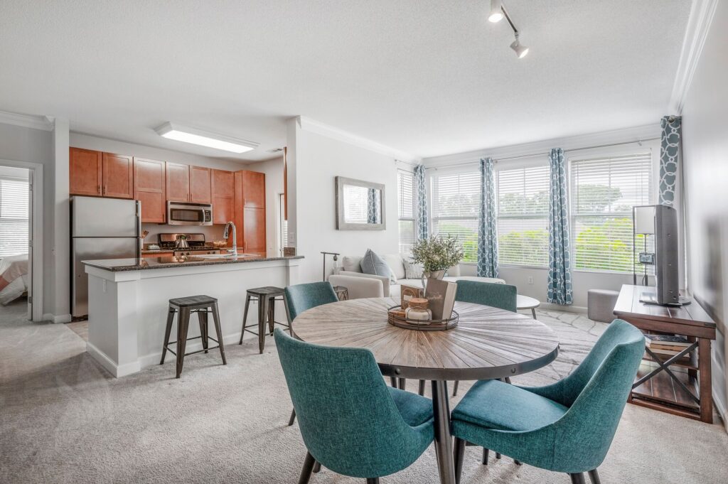 Modern open-plan living space with a kitchen, round dining table with teal chairs, living area with sofa and TV, large windows with curtains, and neutral tones throughout.