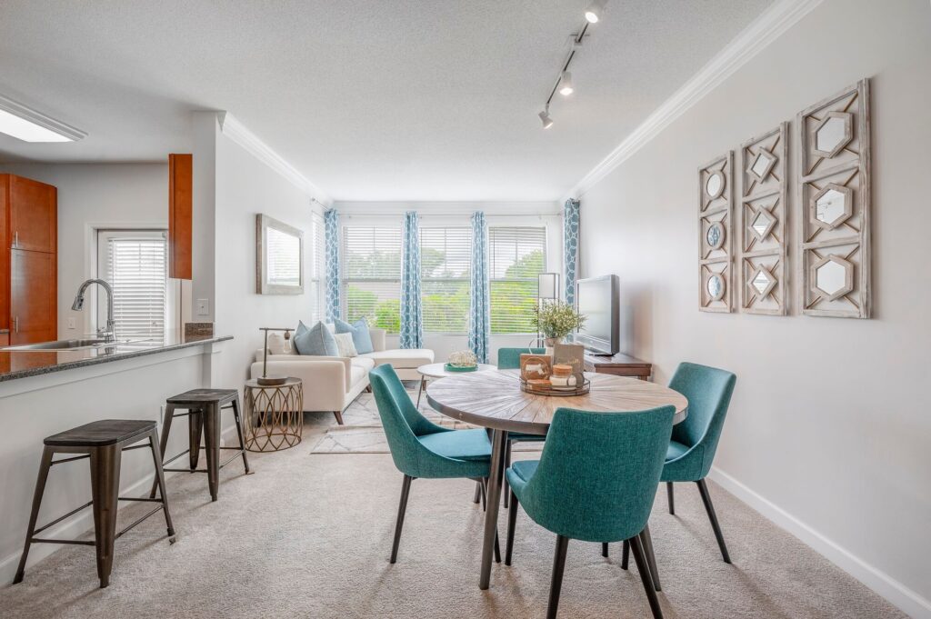 Modern open-concept living and dining area with teal chairs, round table, cozy sofa, large windows with blue curtains, and minimalist decor. Kitchen with bar stools is visible on the left.
