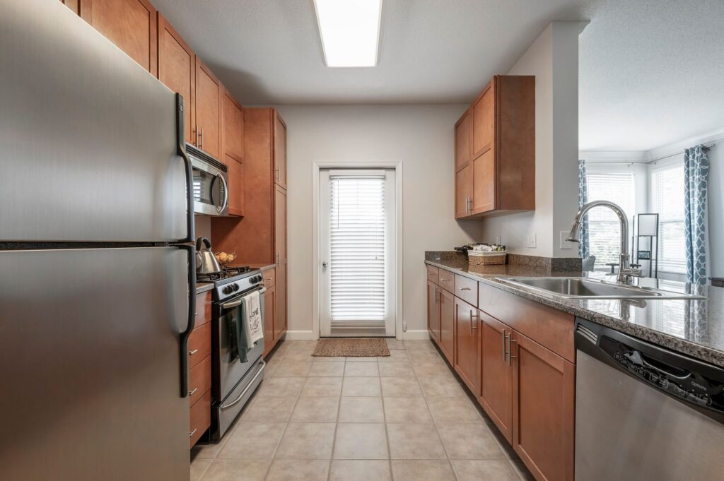 Modern kitchen with stainless steel appliances, wooden cabinets, tile floor, and granite countertops. A door with blinds leads outside. Natural light brightens the space.
