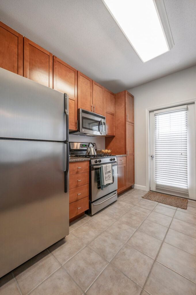 Modern kitchen with stainless steel fridge, oven, and microwave, brown wooden cabinets, tile floor, and a door with blinds leading outside. A towel hangs on the oven that says “Home Sweet Home.”.