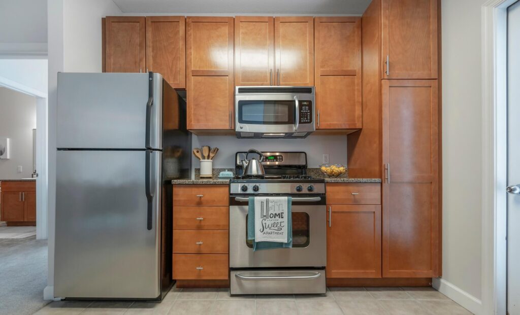 Modern kitchen with stainless steel fridge, oven, and microwave, surrounded by brown wooden cabinets. A kettle, utensil holder, and bowl are on the counter. A towel on the oven reads “Home Sweet Apartment.”.