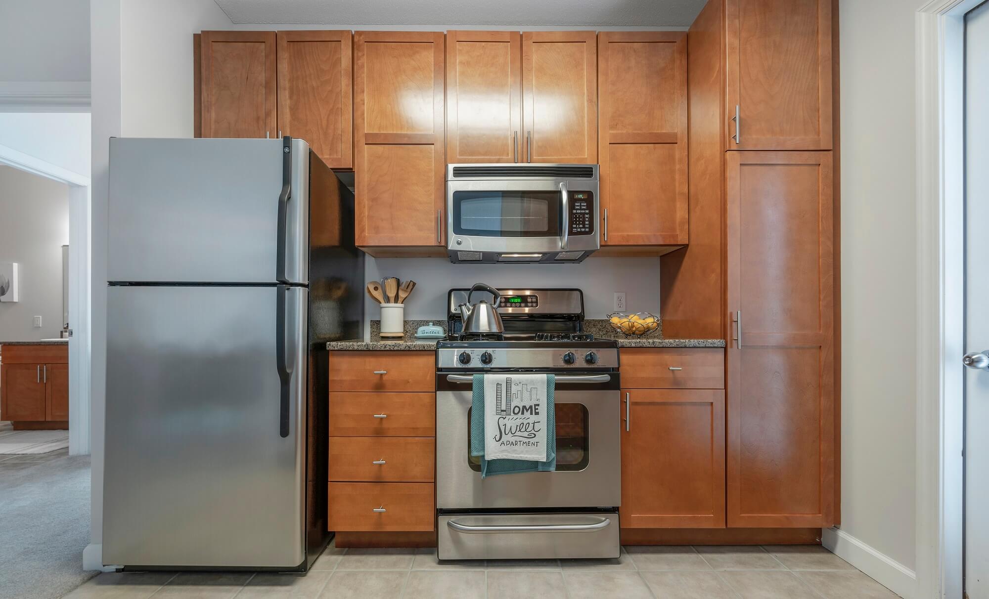 Modern kitchen with stainless steel fridge, oven, and microwave, surrounded by brown wooden cabinets. A kettle, utensil holder, and bowl are on the counter. A towel on the oven reads “Home Sweet Apartment.”.