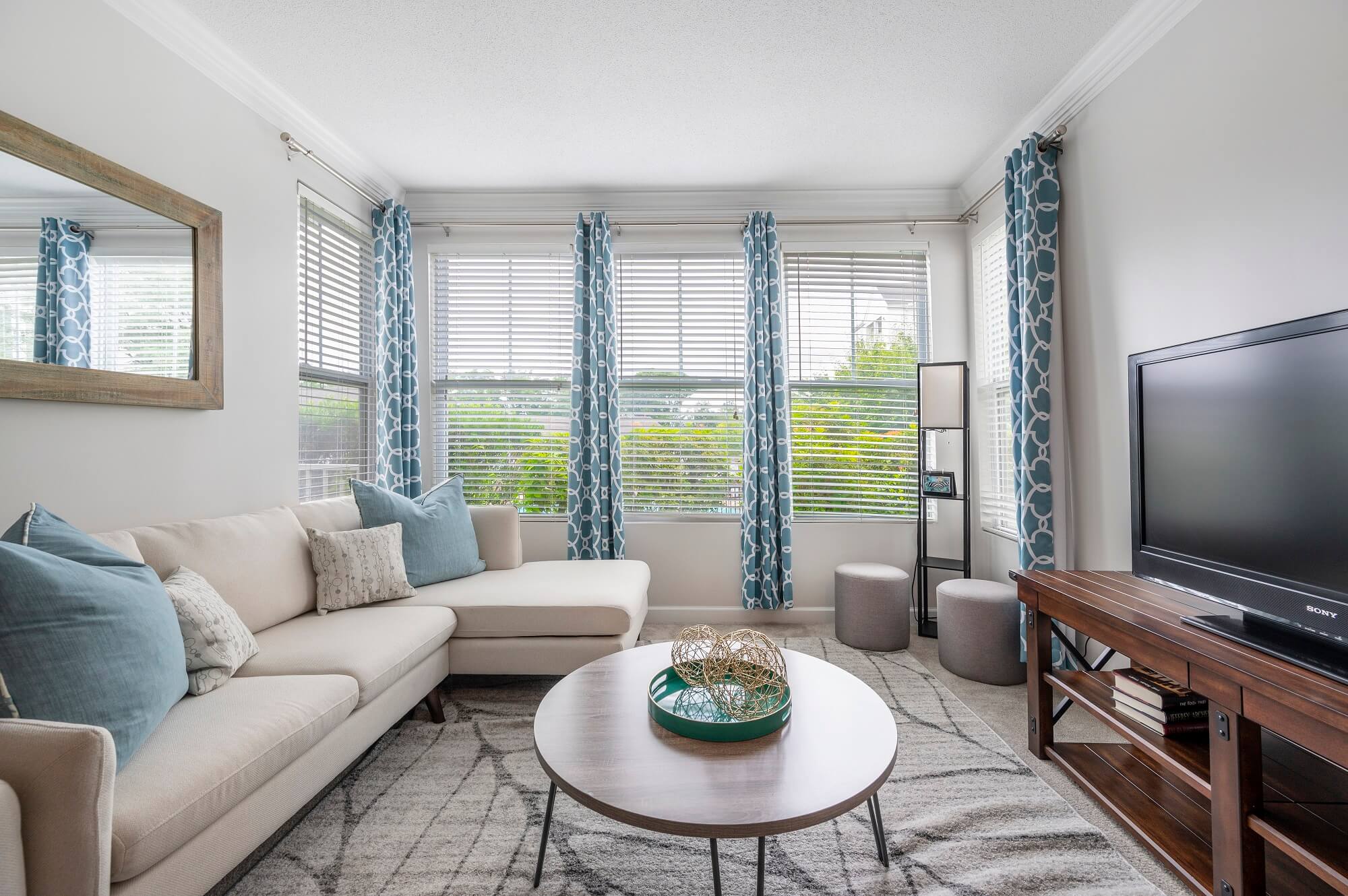 Bright living room with a beige sectional sofa, blue pillows, patterned curtains, a TV on a wooden stand, round coffee table, large windows, and modern decor.