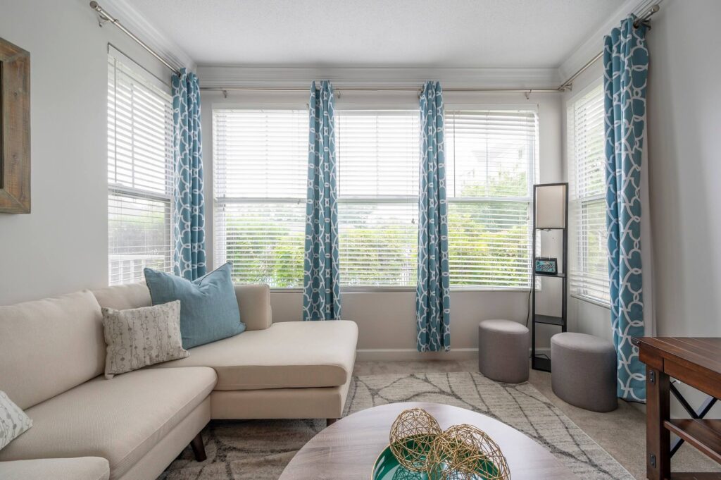 Bright living room with a beige sectional sofa, blue and gray pillows, patterned blue curtains, two gray ottomans, a floor lamp, and a round coffee table with decorative gold spheres. Large windows let in natural light.
