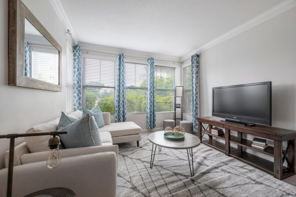 Bright living room with a white sectional sofa, blue pillows, patterned curtains, a round coffee table, TV on a wooden stand, large windows, and a wall mirror. Gray walls and a geometric rug complete the modern decor.