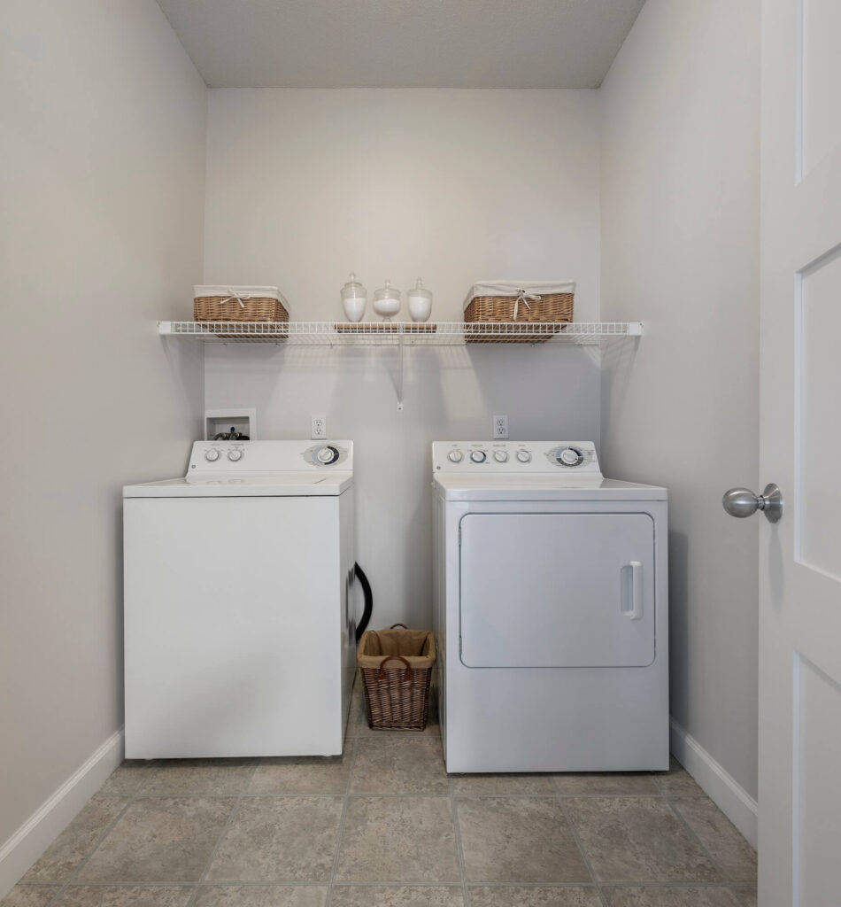 A laundry room with a white washing machine and dryer, a wire shelf above holding baskets and jars, and a small basket on the floor between the appliances. The walls and door are white, and the floor is tiled.