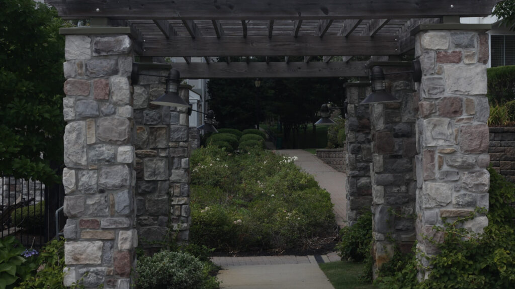 A stone pergola with two hanging lights stands over a garden pathway, surrounded by greenery and shrubs, leading to a sidewalk in a residential area.