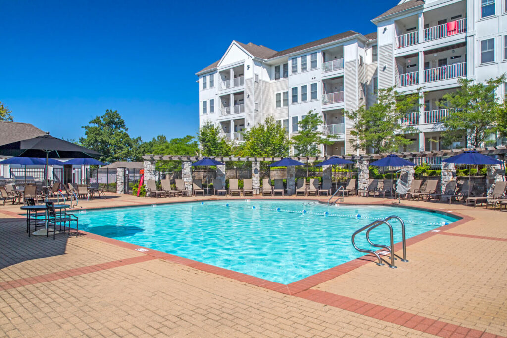 Outdoor swimming pool surrounded by lounge chairs and blue umbrellas, with a multi-story white apartment building and green trees in the background under a clear blue sky.