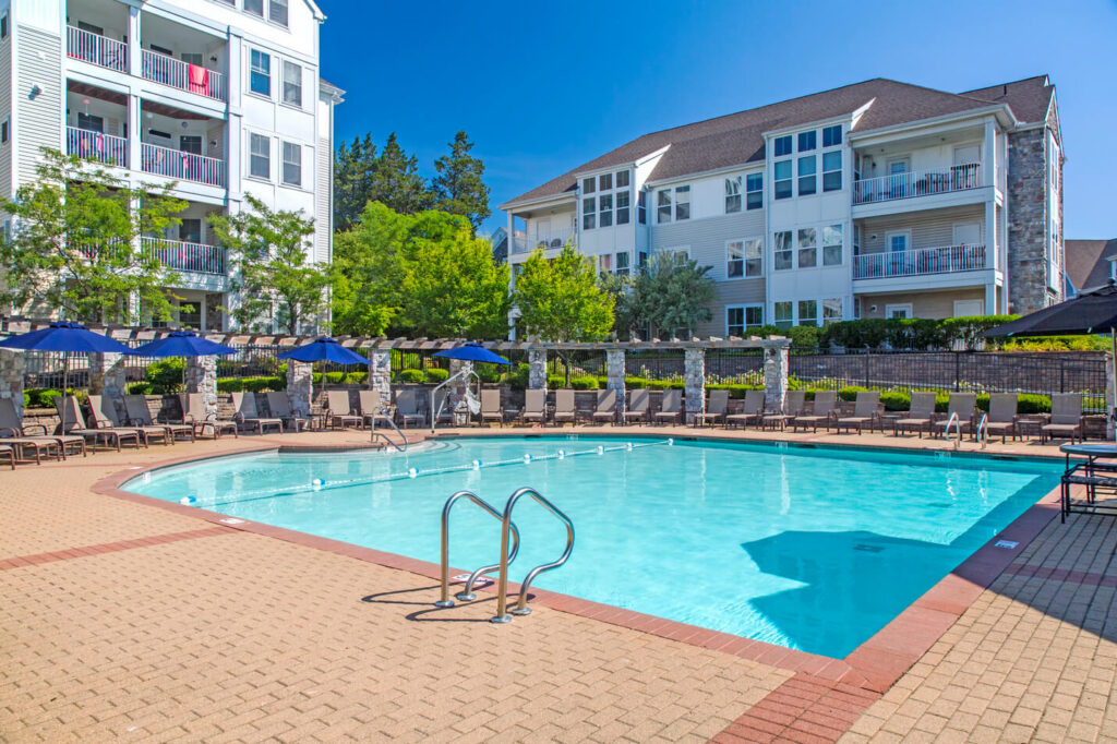 Outdoor swimming pool with metal steps, surrounded by lounge chairs and blue umbrellas, set in front of white apartment buildings with balconies and greenery under a clear blue sky.