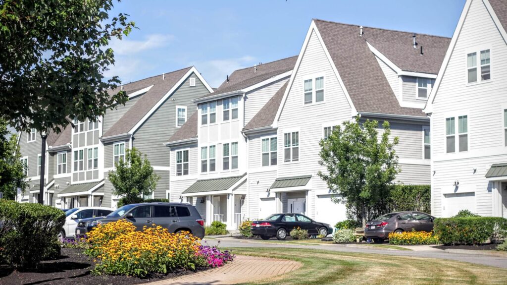 Row of modern, three-story townhouses with white siding and peaked roofs, cars parked in front, and colorful flowers and greenery along a curved walkway on a sunny day.