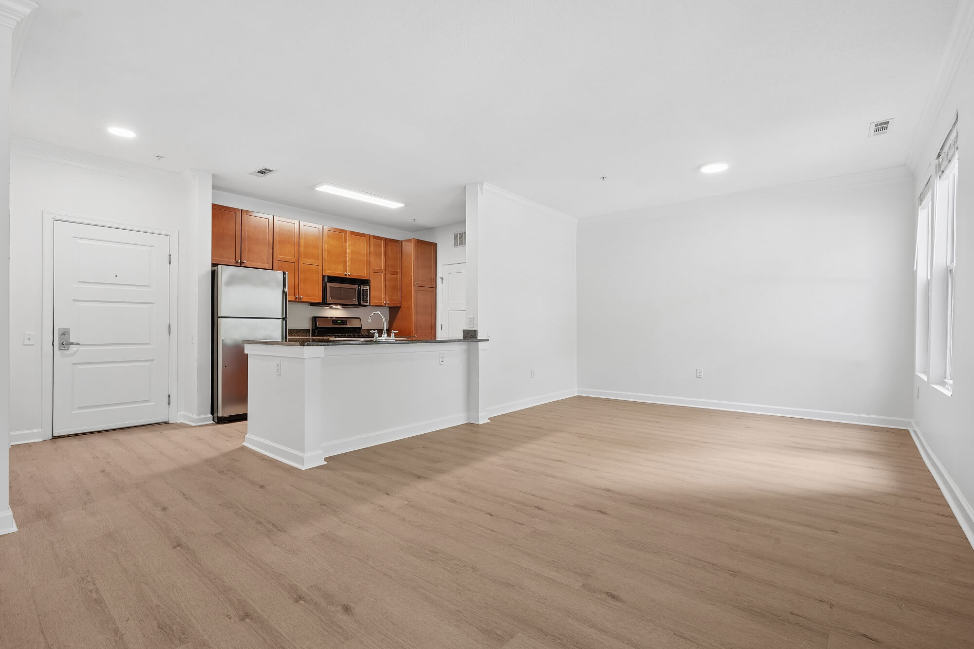 An open-plan apartment featuring a kitchen with wood cabinets, stainless steel appliances, and a peninsula counter overlooking a spacious, empty living area with light-toned wood flooring.