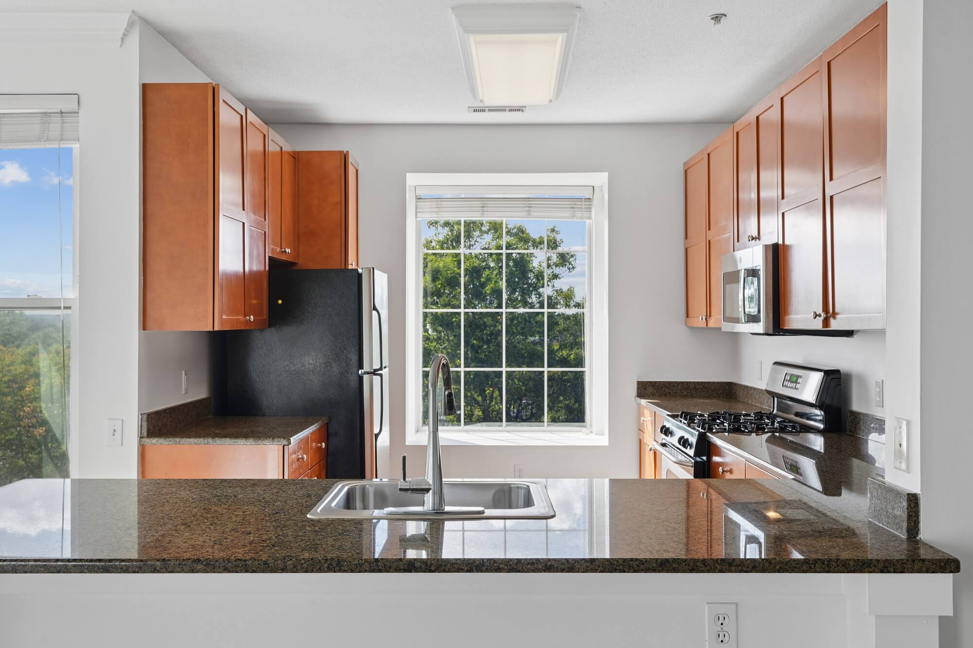 A kitchen featuring brown wood cabinets, a black refrigerator, a granite countertop with a sink, and a large window looking out onto trees.